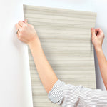 A person wearing a white and black striped shirt is applying York Wallcoverings' Pandora Leaf Oak Wallpaper Brown (60 Sq.Ft.), featuring beige and white stripes, to a wall. They use both hands to smooth and align the wallpaper for easy installation and removal. A silver ring gleams on one of the person's fingers.