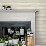 A fireplace adorned with an array of potted succulents in white ceramic planters is set against a backdrop of Pandora Leaf Oak Wallpaper Brown (60 Sq.Ft.) by York Wallcoverings. The mantle and hearth are filled with various succulent plants, creating a lush, green display. A small wooden stool beside the fireplace also holds a few potted succulents.