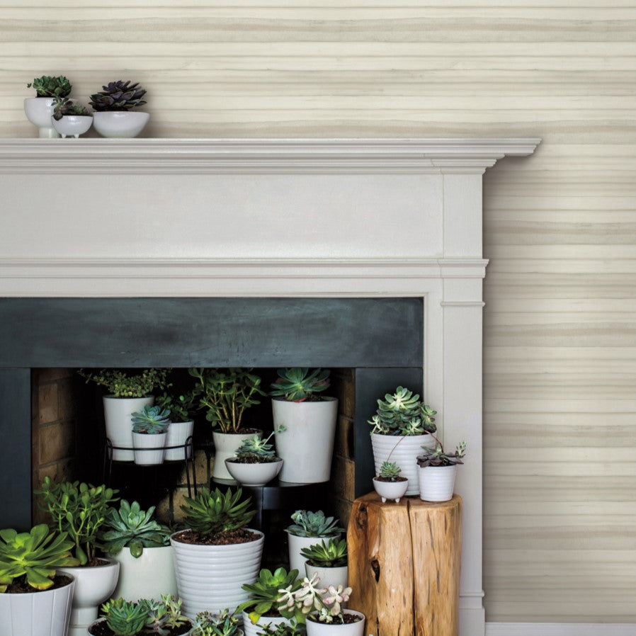 A fireplace adorned with an array of potted succulents in white ceramic planters is set against a backdrop of Pandora Leaf Oak Wallpaper Brown (60 Sq.Ft.) by York Wallcoverings. The mantle and hearth are filled with various succulent plants, creating a lush, green display. A small wooden stool beside the fireplace also holds a few potted succulents.