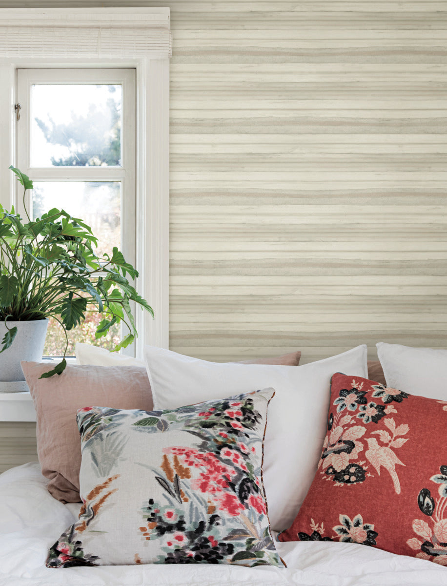 A cozy bed is adorned with white, grey, and floral-patterned pillows in front of a large window. A vibrant green potted plant sits on the windowsill, and the room features light-colored striped Pandora Leaf Oak Wallpaper Brown (60 Sq.Ft.) by York Wallcoverings with tropical foliage. Sunlight streams in through the window, adding a warm touch.