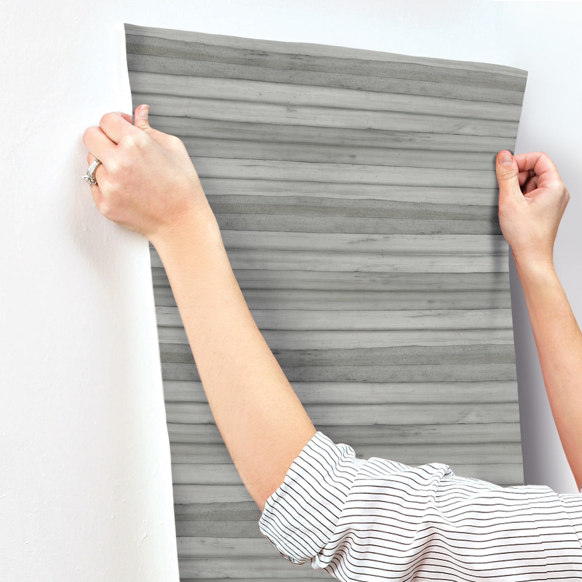 A person wearing a white and black striped shirt is applying a sheet of gray, horizontally striped Pandora Leaf Oak Wallpaper Brown (60 Sq.Ft.) by York Wallcoverings to a white wall. They're holding the top corners to ensure it's aligned properly, showcasing how easy installation and removal can be. A ring is visible on their left hand.