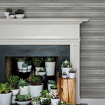 A white fireplace mantel with a black interior opening is filled with an assortment of potted succulents in white and light-colored pots. A wooden stump holds additional succulents to the right side of the fireplace. The background, featuring Pandora Leaf Oak Wallpaper Brown (60 Sq.Ft.) by York Wallcoverings, showcases tropical foliage.