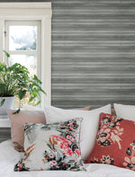 A cozy bedroom scene featuring a bed with multiple pillows in floral patterns and solid colors. Above the bed, there is a window with an abundance of natural light, and a potted plant with tropical foliage on the windowsill. The walls are adorned with York Wallcoverings' Pandora Leaf Oak Wallpaper Brown (60 Sq.Ft.) for easy installation and removal.