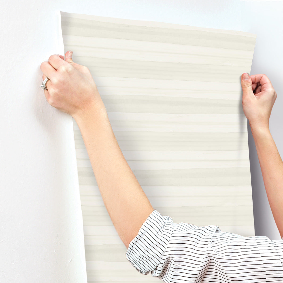 A person with a striped shirt is hanging a piece of neutral-colored, horizontally-striped Pandora Leaf Oak Wallpaper Brown (60 Sq.Ft.) by York Wallcoverings onto a white wall. Their hands are holding the top corners of the wallpaper, ensuring it is aligned properly. A ring is visible on one of their fingers.