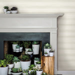 A collection of potted succulents is arranged on a fireplace mantle and inside the fireplace opening. The soft beige wall, with its York Wallcoverings Pandora Leaf Oak Wallpaper Brown (60 Sq.Ft.), and monochromatic plant pots add to the serene and modern aesthetic of the scene. A tree stump sits beside the fireplace with more succulents on it.