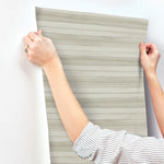 A person wearing a striped shirt is seen adjusting a piece of Pandora Leaf Oak Wallpaper Brown (60 Sq.Ft.) by York Wallcoverings on a white wall. The hands are holding the top edges of the wallpaper, which features horizontal stripes in varying shades of gray, showcasing its easy installation and removal.