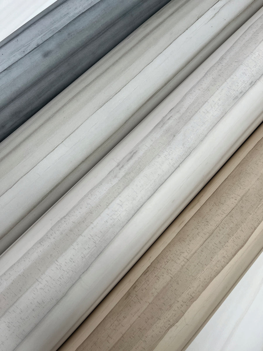 A close-up image of several rolls of fabric in varying shades of grey, white, and beige, laid out diagonally. The textures appear smooth, and the fabrics are neatly arranged to display the different hues and tones, reminiscent of York Wallcoverings' Pandora Leaf Oak Wallpaper Brown (60 Sq.Ft.) easy installation and removal.