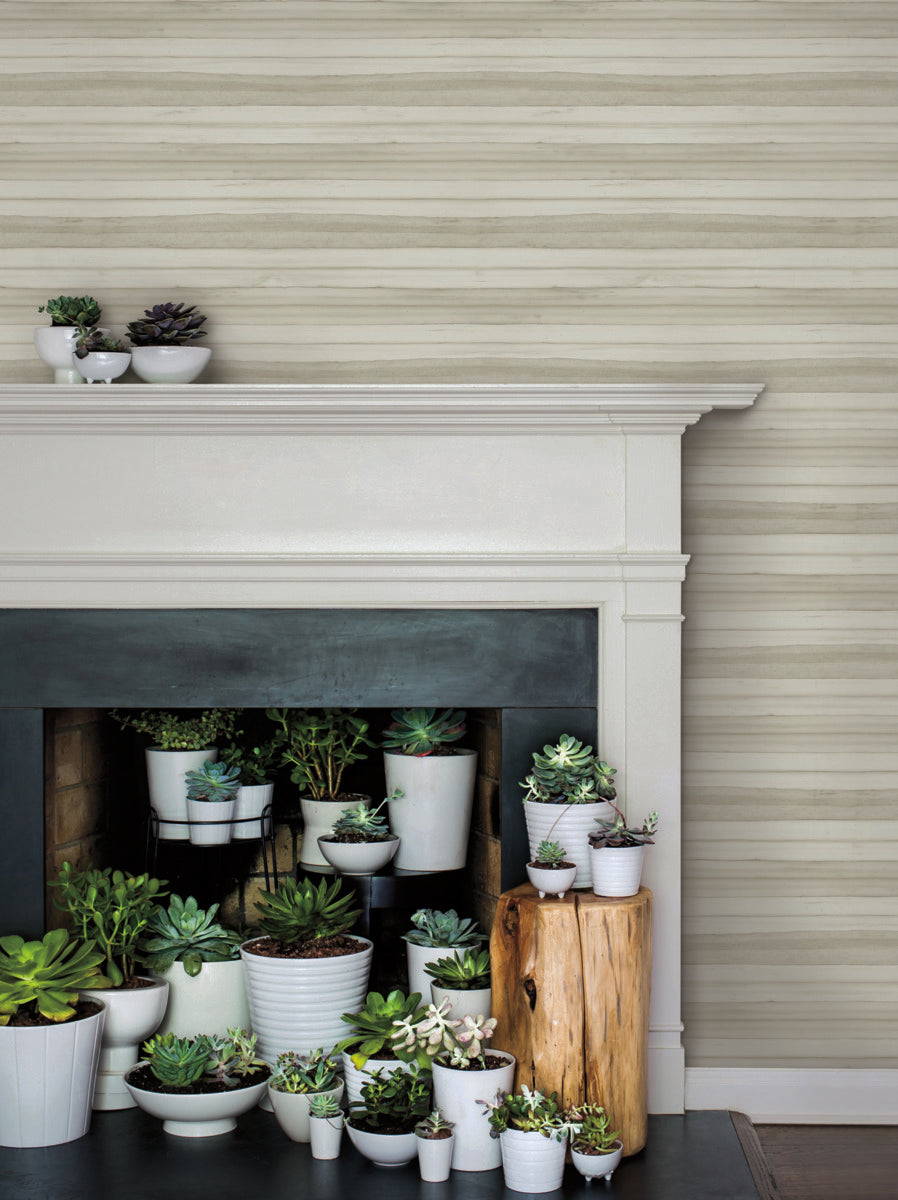 A fireplace mantle decorated with various small potted succulents in white pots. Below the mantle, more succulents are arranged inside the non-burning fireplace and on two wooden stump stools to the right. The wall features light horizontal stripes, easily transformed with York Wallcoverings' Pandora Leaf Oak Wallpaper Brown (60 Sq.Ft.).