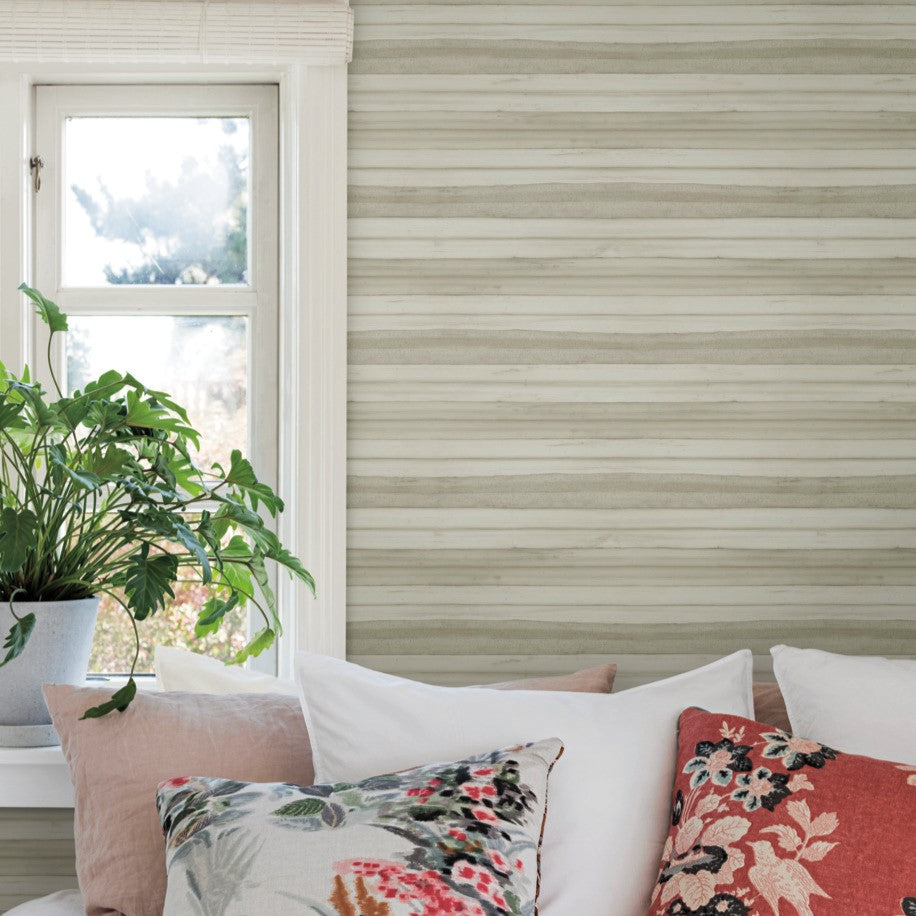 A cozy bedroom setup with York Wallcoverings' Pandora Leaf Oak Wallpaper Brown (60 Sq.Ft.) as a chic backdrop. The bed features white and floral-patterned pillows. A large potted plant with tropical foliage sits on a wooden side table near a window that lets in natural light, enhancing the serene atmosphere.