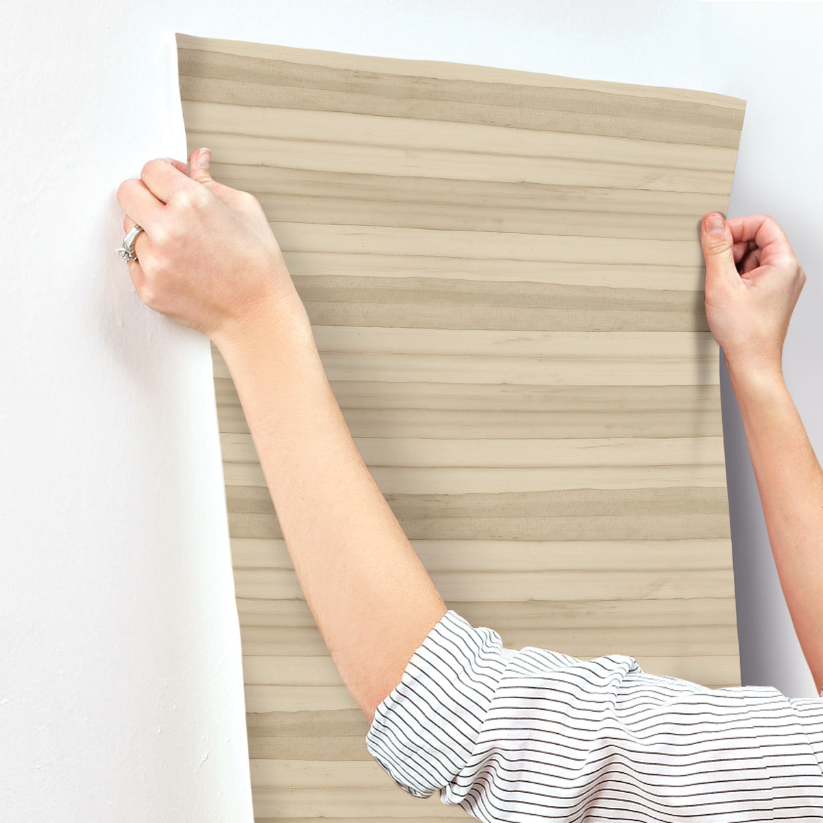 A person is applying a sheet of Pandora Leaf Cotton Wallpaper White/Off White (60 Sq.Ft.) by York Wallcoverings with a light wood grain pattern onto a white wall. The person's hands are holding up the SureStrip™ paper, and they are wearing a white shirt with black stripes and a ring on one finger.