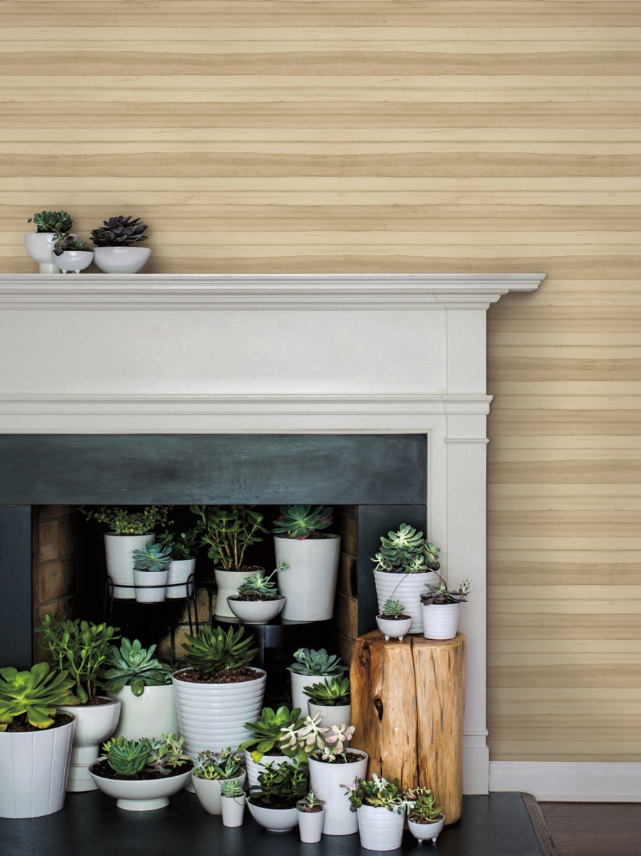 A fireplace is filled with various potted succulents and small plants, arranged neatly in white pots of different sizes. A rustic wooden log adds a decorative element, and more succulent pots are placed on the white mantel above. The wall has a beige and brown striped pattern, enhanced by York Wallcoverings Pandora Leaf Oak Wallpaper Brown (60 Sq.Ft.) for an easy installation and removal.