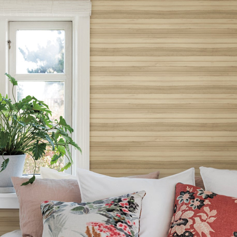 A small window with white trim is set in a wood-paneled wall adorned with Pandora Leaf Oak Wallpaper Brown (60 Sq.Ft.) by York Wallcoverings. A plant in a gray pot sits on the windowsill to the left. Below, a couch features various patterned pillows in white, red, and floral designs.