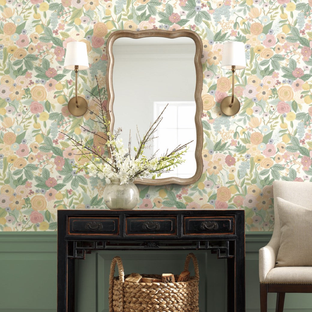 The room's striking design, akin to York's Pastels Garden Party Wallpaper, showcases a mirror with a wooden frame and two wall sconces. Beneath them, a dark wooden console table supports a vase of branches and a woven basket. A beige armchair is partially visible on the right side.