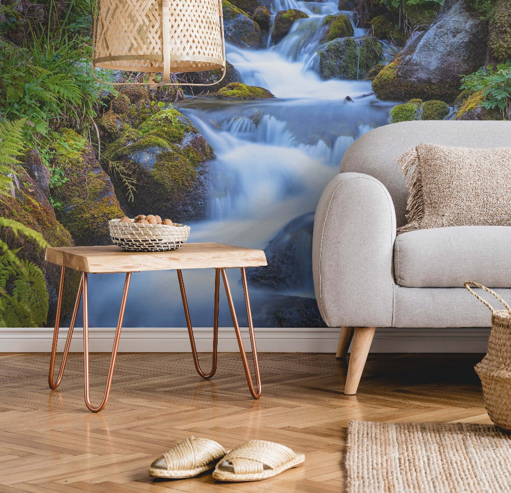 A cozy living room corner with a modern beige sofa, a wooden coffee table with copper legs, and a large Decor2Go Wallpaper Mural of a serene Peaceful Waterfall surrounded by greenery. A pair of woven slippers rests