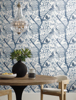 A round wooden table adorned with a black vase containing green foliage, a small pitcher, and a bowl of lemons sits under a modern glass chandelier. The background showcases Peacock Block Print Wallpaper in blue by York Wallcoverings, which features blue and white botanical patterns with branches and leaves designed from durable materials for a nature-inspired aesthetic.