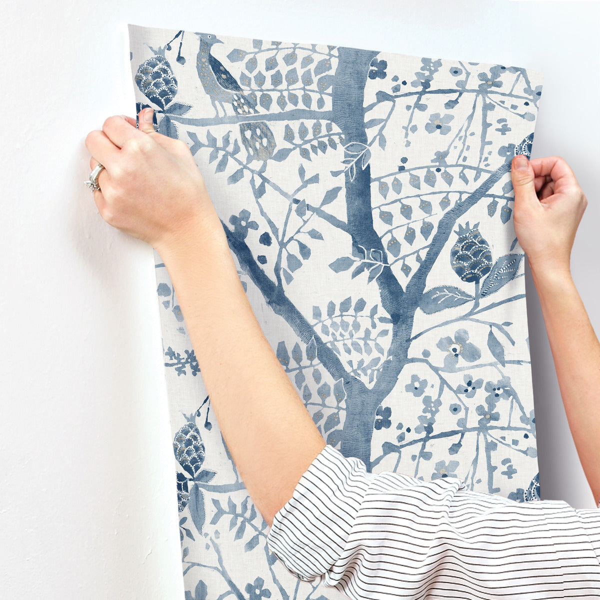 A person with rolled-up sleeves hangs the Peacock Block Print Wallpaper Blue by York Wallcoverings, which features a nature-inspired blue and white botanical pattern of branches, leaves, and flowers. The person's hands carefully adjust the durable wallpaper on a white wall.