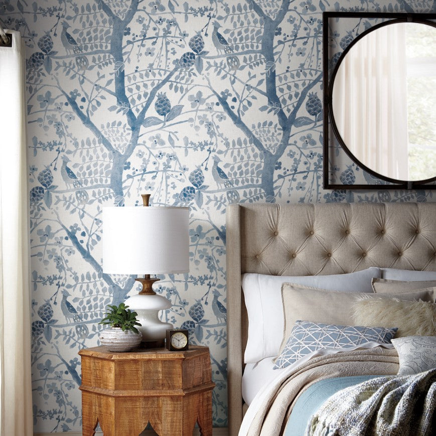 A cozy bedroom showcasing a bed with white and beige bedding, a tufted headboard, and a round mirror on the wall. A wooden nightstand supports a white lamp, a plant, and a clock. The York Wallcoverings Peacock Block Print Wallpaper in Blue (60 SqFt) adds an elegant touch to the nature-inspired design.