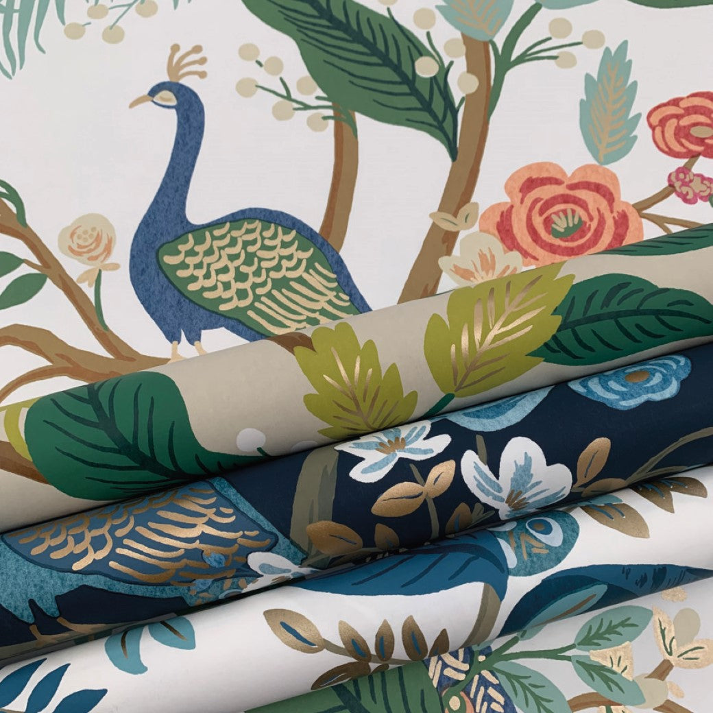 The Periwinkle Peacock Wallpaper by York offers a close-up view of colorful fabric rolls with a nature theme, featuring peacocks, flowers, and branches in shades of blue, green, and red. Its vibrant Chinoiserie design is reminiscent of the decorative patterns by Rifle Paper Co.