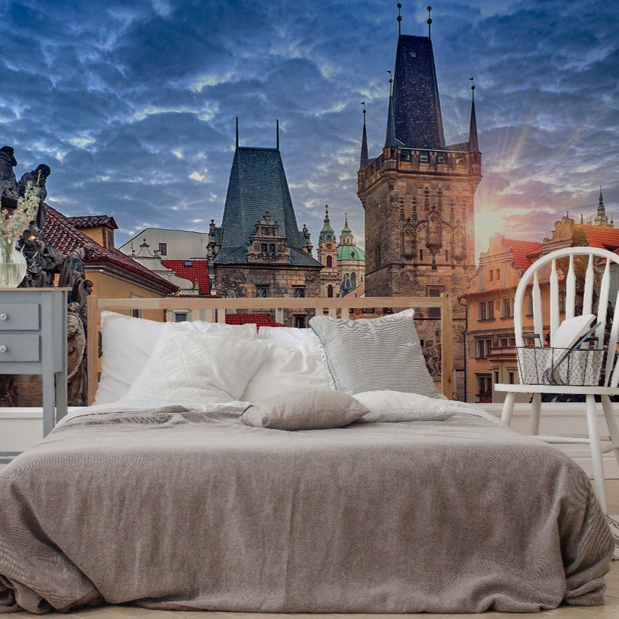 A bed with white and grey bedding is set against a surreal backdrop of Prague Alley Wallpaper Mural by Decor2Go Wallpaper Mural, featuring iconic spires under a dramatic sunrise sky.