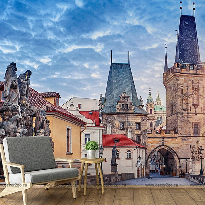 A vivid mural of Charles Bridge and Prague's skyline on a wall in a room with a modern armchair and a small table, blending indoor comfort with iconic outdoor scenery through Decor2Go Wallpaper Mural.