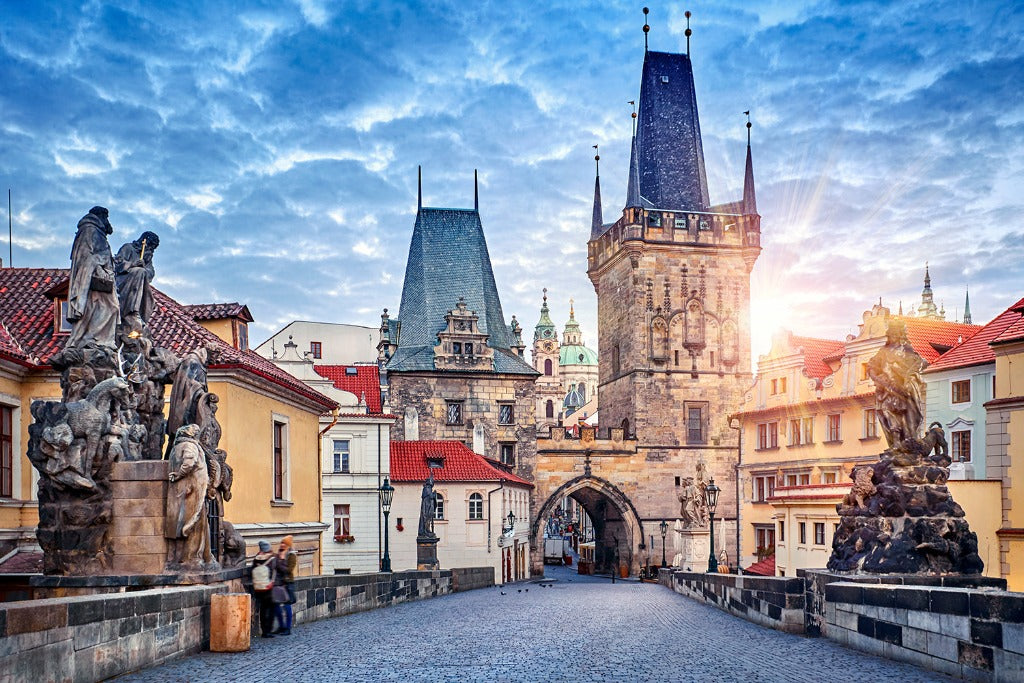 Sunrise over Charles Bridge in Prague, showcasing the historic towers and statues with vibrant skies and scattered clouds, as a few people wander the cobblestone path on their Decor2Go Wallpaper Mural.