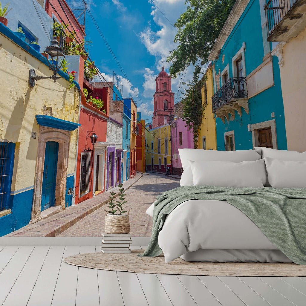 A surreal image blending a cozy bedroom setting with an outdoor vibrant street scene featuring colorful buildings and a clear blue sky. A bed is positioned in the foreground on white flooring, accented by Decor2Go Wallpaper Mural.