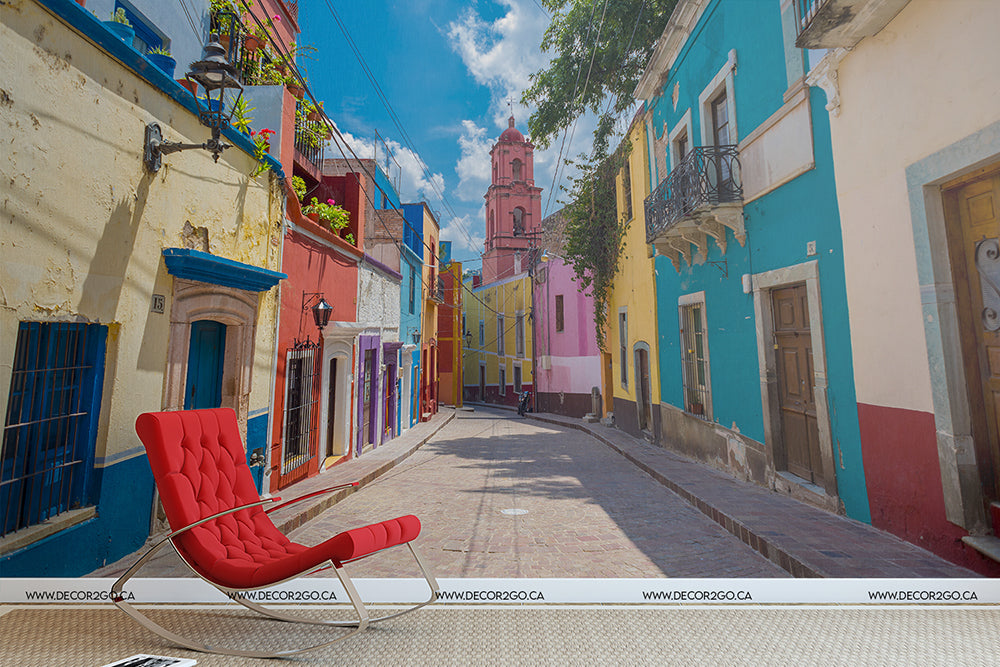A vibrant, colorful street features rows of pastel-colored buildings under a bright blue sky. A modern red lounge chair sits prominently in the foreground of the cobbled street, perfectly complementing the Decor2Go Wallpaper Mural Rainbow Road Wallpaper Mural.