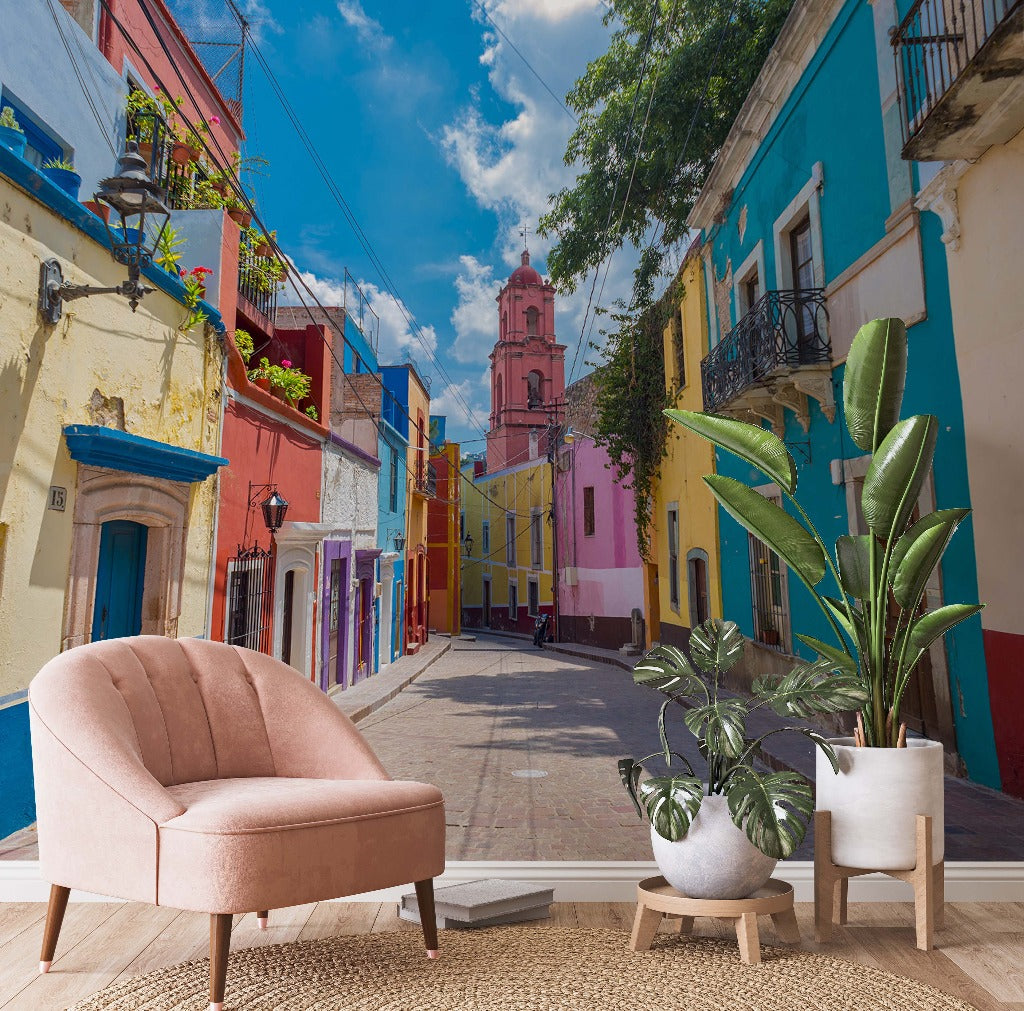 A vibrant street with colorful buildings and a distant pink church tower, seamlessly blending into a room with Decor2Go Wallpaper Mural's Rainbow Road wallpaper and potted plants, creating an indoor-outdoor illusion.