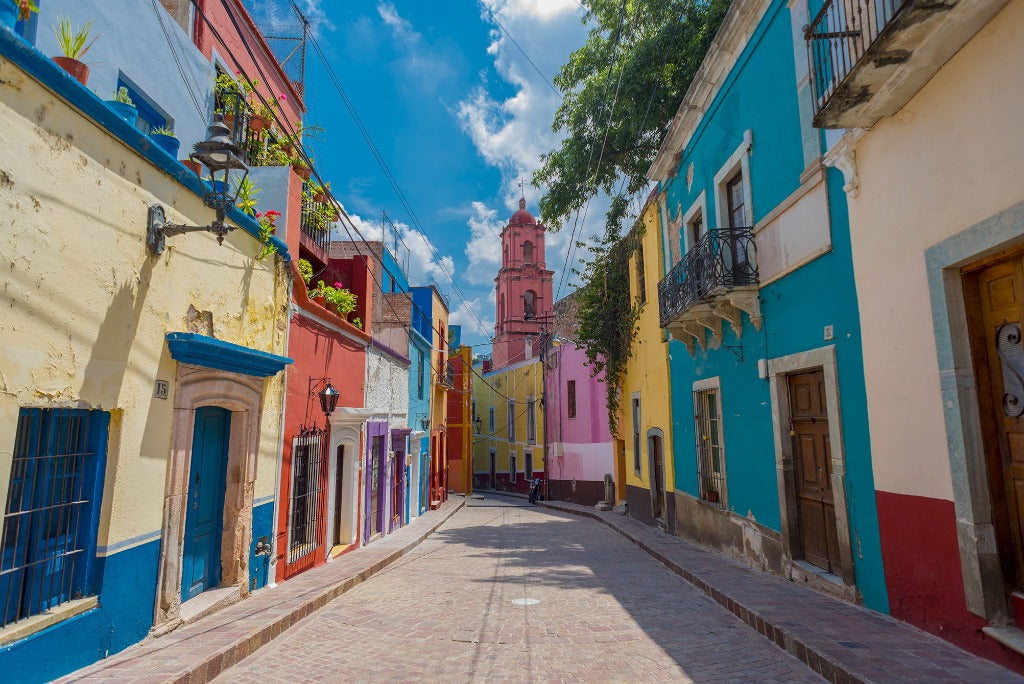A vibrant street, reminiscent of Decor2Go Wallpaper Mural, is lined with colorful colonial-style buildings under a clear blue sky, leading towards a pink church tower in the distance.