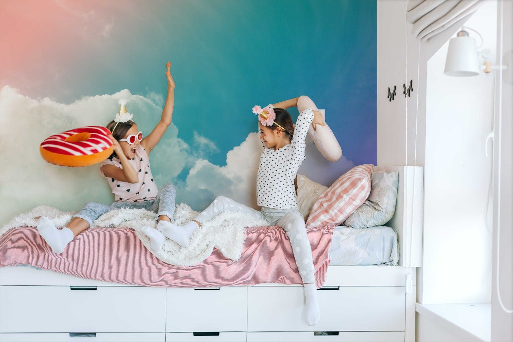Two young children having a playful pillow fight on a bed, surrounded by fluffy cloud decorations, against a Rainbow Sky Wallpaper Mural from Decor2Go Wallpaper Mural. They wear crowns and whimsical masks.