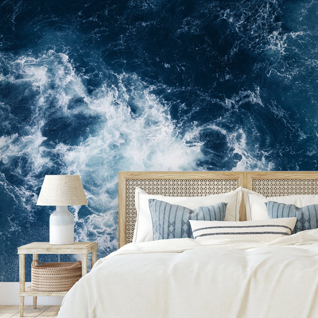 A modern bedroom featuring a queen-sized bed with white and blue bedding between two side tables, each with a lamp, against a Decor2Go Wallpaper Mural of Realm of Poseidon swirling blue ocean waves.