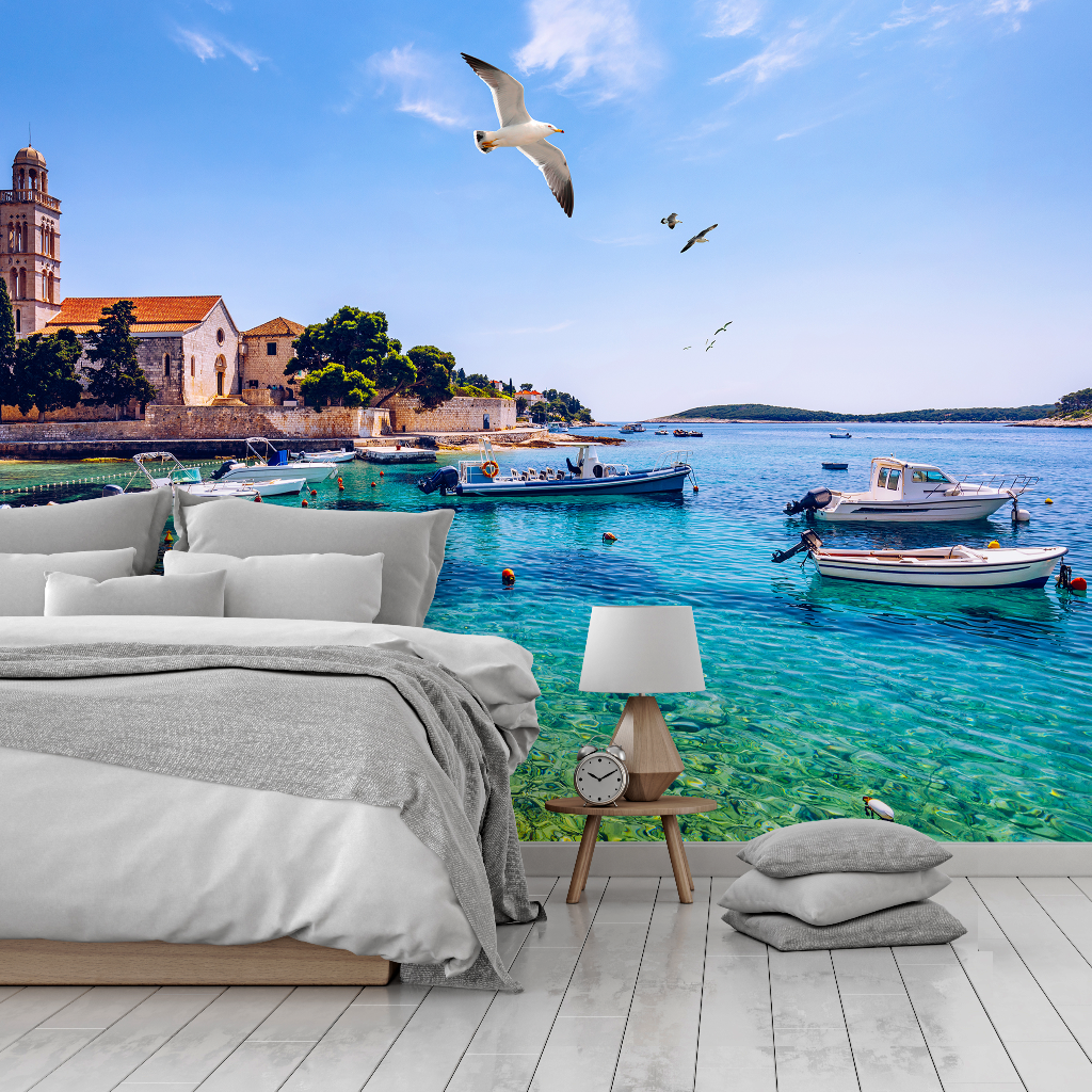 A bedroom with an immersive Decor2Go Wallpaper Mural of a scenic coastline featuring boats on clear blue water, a historic building in the background, and seagulls flying overhead.