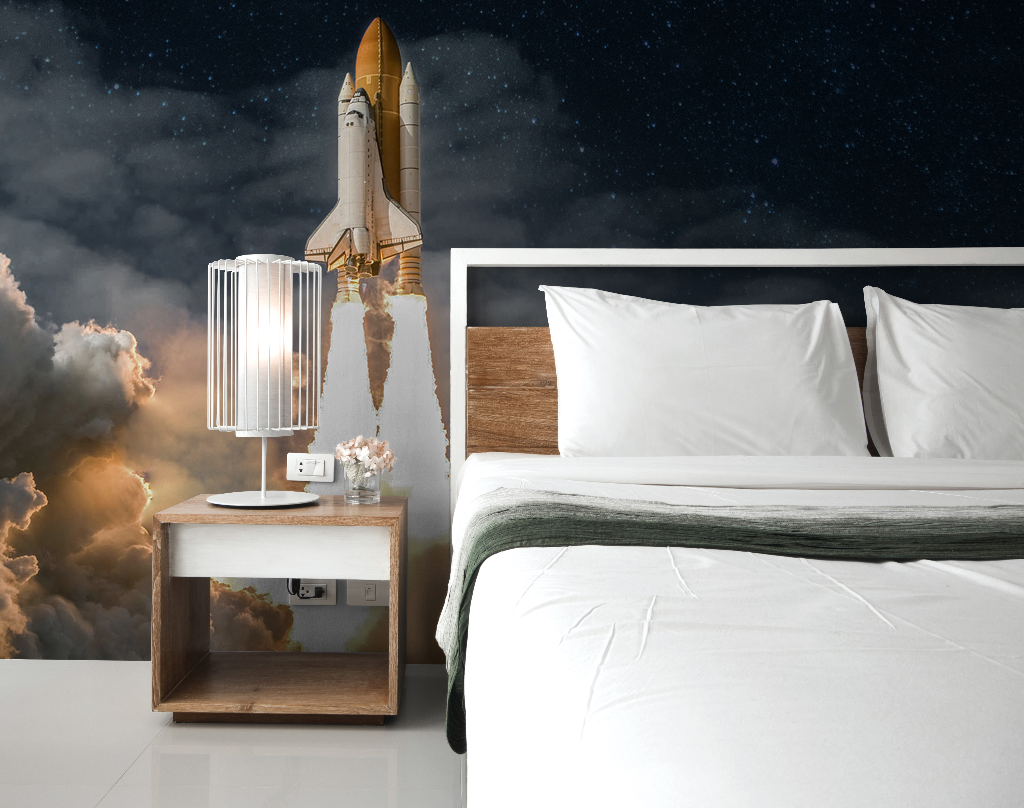 A surreal bedroom with a Decor2Go Wallpaper Mural featuring a rocket launching into a starry night sky, next to a modern bed with white bedding and a stylish bedside lamp.