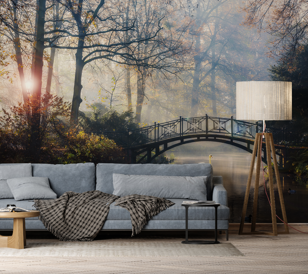 A cozy living room featuring a gray sofa with a plaid throw, a floor lamp, and a side table, with a Decor2Go Wallpaper Mural of the Romantic Bridge in autumn on the wall.