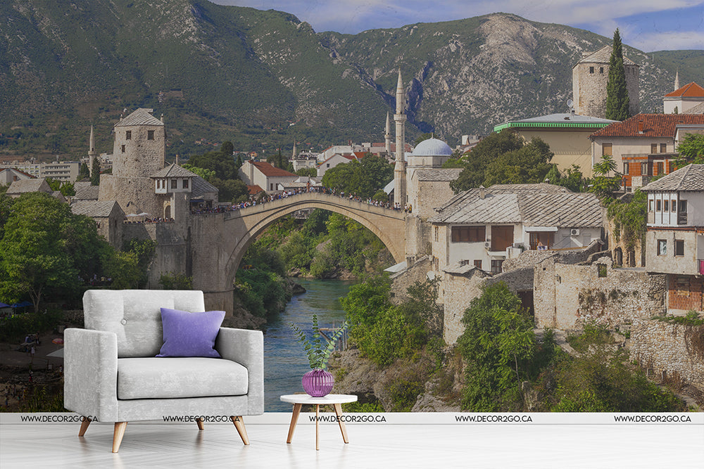 A surreal composition blending an indoor scene with an outdoor historical landscape, featuring a white armchair and side table set against a Decor2Go Wallpaper Mural depicting the Stari Most bridge and scenic town in Mostar.