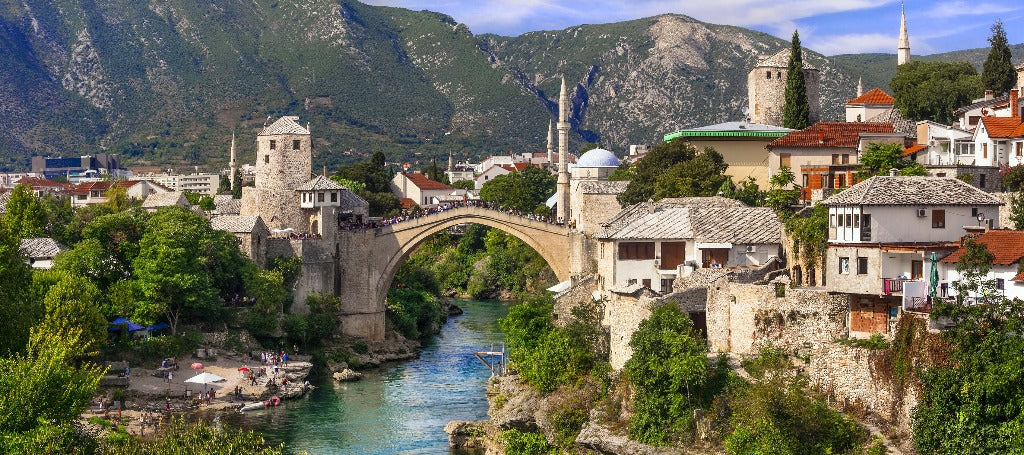 Historic stone bridge arching over a river in a lush valley with traditional buildings and green mountains in the background, perfect for Decor2Go Wallpaper Mural.