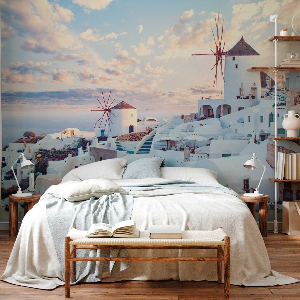 A cozy bedroom with a large bed covered in a beige duvet, a wooden bench with an open book at the foot, and a wall-sized Decor2Go Wallpaper Mural depicting its iconic Santorini Skyline.