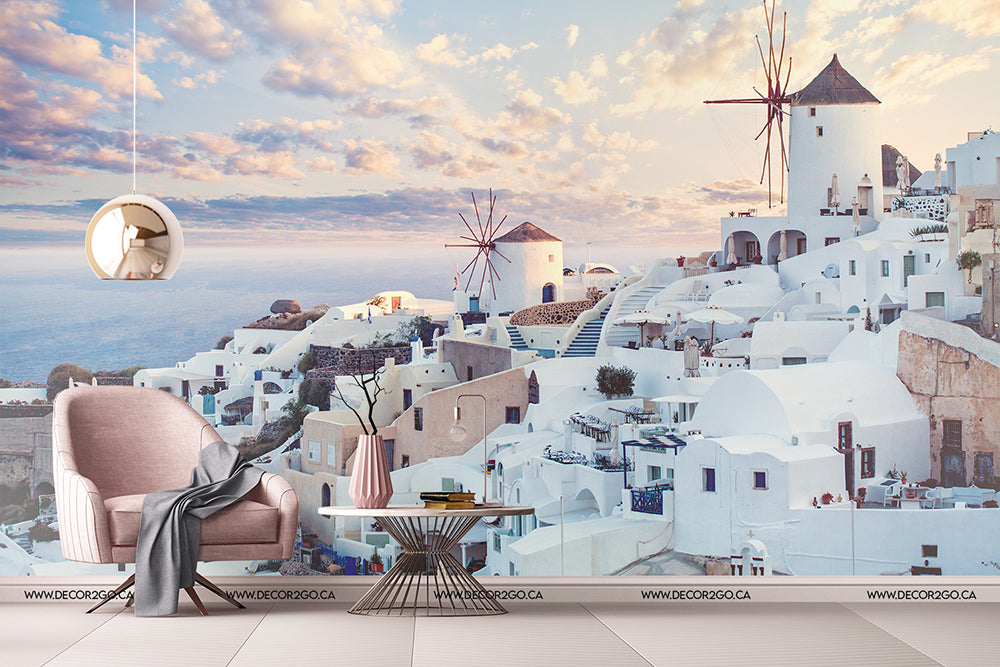 A surreal room blends into a scenic view of Santorini, Greece, with iconic white buildings and windmills captured in the Decor2Go Wallpaper Mural. A stylish chair and table are set.