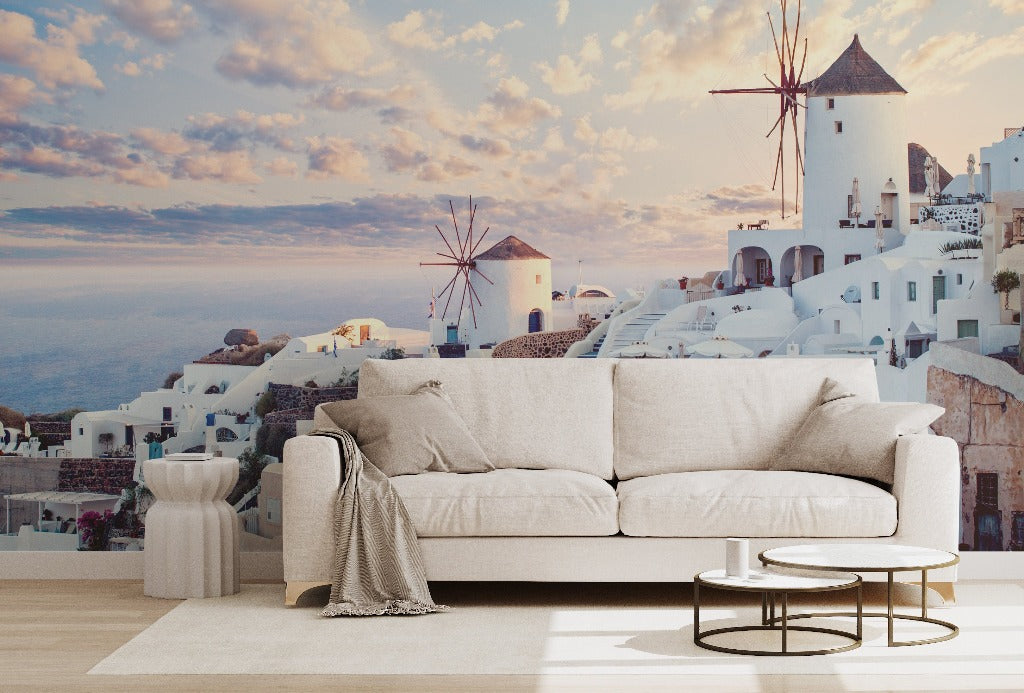 A modern living room with a white sofa and small tables in front of a large window displaying a Decor2Go Wallpaper Mural, showing its famous white houses and windmills against a sunset sky.
