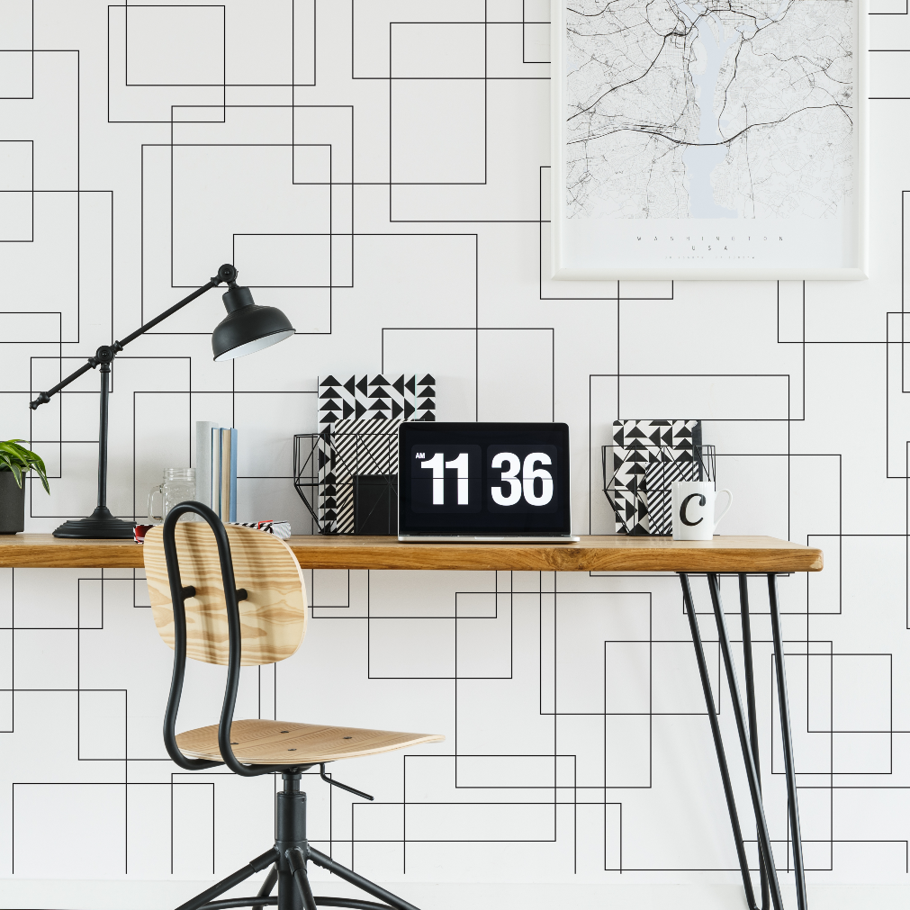 A modern home office with a sleek wooden desk against a Decor2Go Wallpaper Mural. The desk displays a digital clock, striped notebooks, a coffee mug, and an adjustable lamp. A stylish chair and