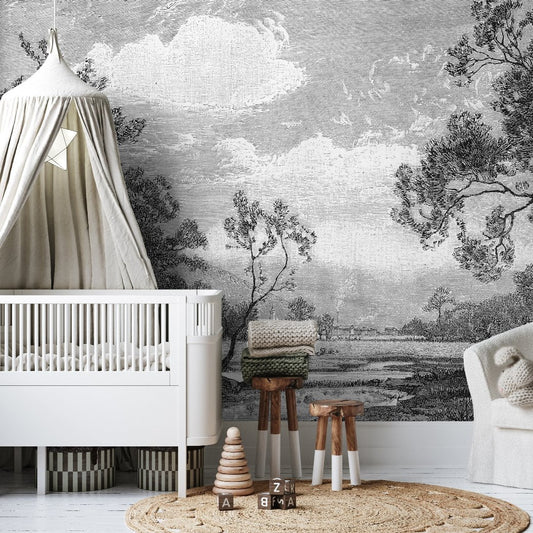 A serene nursery features a white crib with a draped canopy, a soft armchair, and a stool with folded blankets. Wooden stacking toys and alphabet blocks adorn a light-colored rug. The backdrop is the Serene Forest Wallpaper Mural by Decor2Go Winnipeg, depicting trees and clouds, creating an aura of timeless elegance.