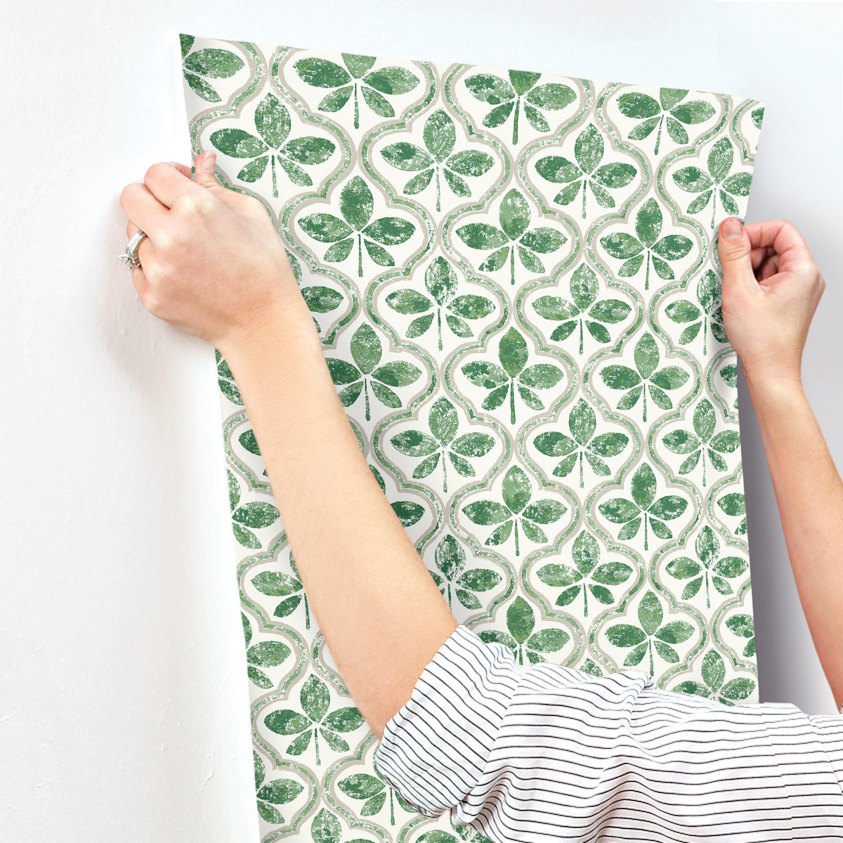 A person with rolled-up sleeves is seen applying Sevilla River Rock Wallpaper Grey (60 Sq.Ft.) by York Wallcoverings with a repeating green shamrock pattern on a white background to a wall. The wallpaper has a vintage, slightly distressed look. The person's hands are carefully aligning the paper for application, showcasing geometric elegance.