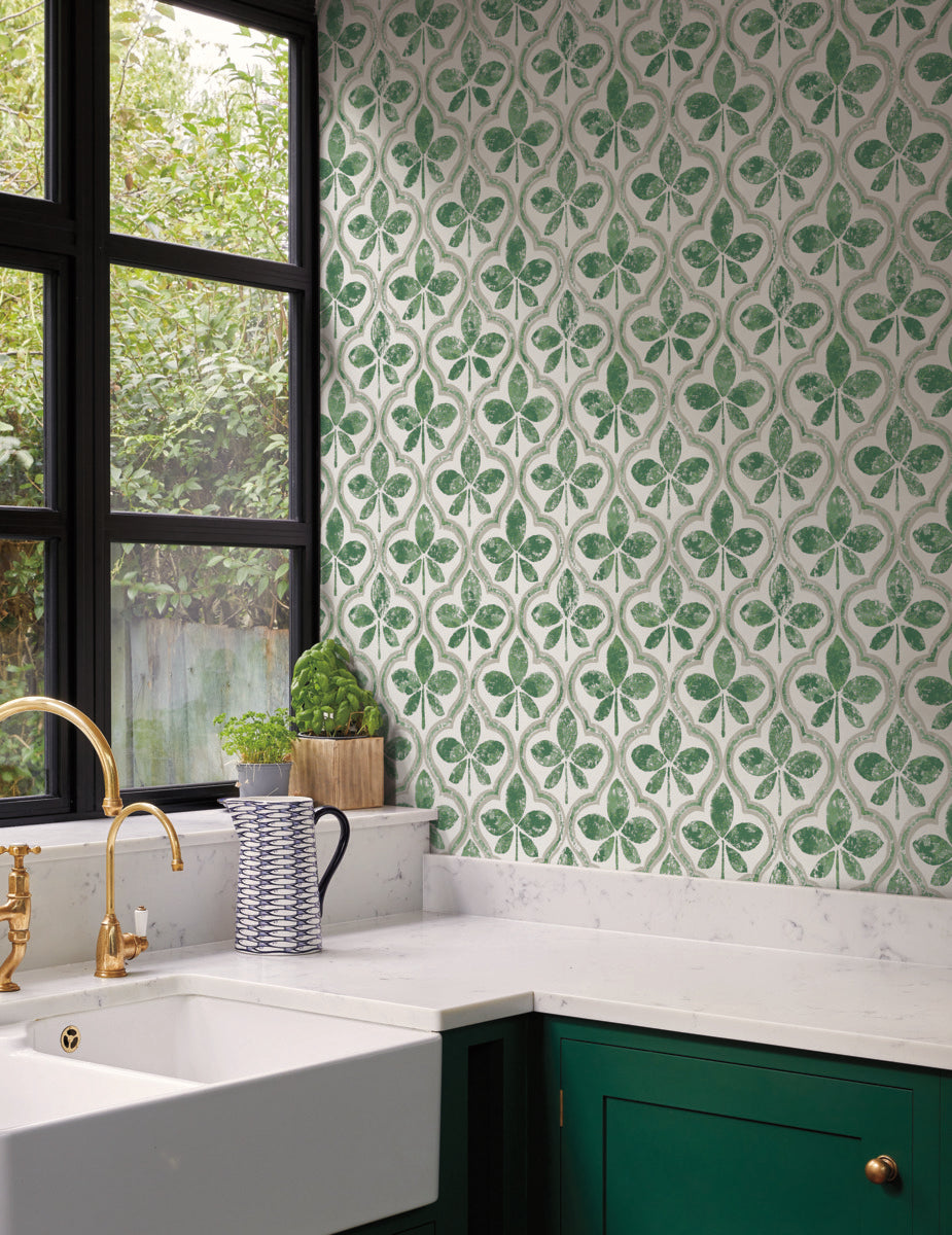 A modern kitchen corner features a large white farmhouse sink, a gold faucet, and green cabinets. The countertop holds a white and blue pitcher and a small plant. The walls display York Wallcoverings Sevilla Onyx Wallpaper Black, Brown (60 Sq.Ft.) with green and white patterns, and there is a large window providing natural light.