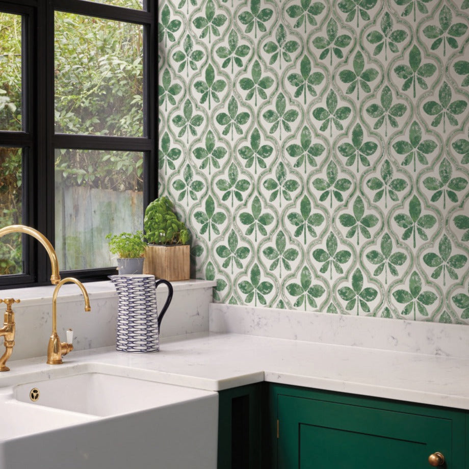 A kitchen features green Sevilla Clover Wallpaper Green (60 Sq.Ft.) by York Wallcoverings with a geometric leaf design, green lower cabinets, a marble countertop, and a white farmhouse sink with a brass faucet. A potted plant and ceramic pitcher near the window allow light to fill the space.