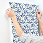 A person is hanging Sevilla River Rock Wallpaper Grey (60 Sq.Ft.) by York Wallcoverings with a floral trefoil pattern in blue and white. They are wearing a striped shirt, and their arms are partially visible, aligning the wallpaper against a white wall. The design showcases symmetrical, repeating motifs printed on SureStrip Substrate for easy application.