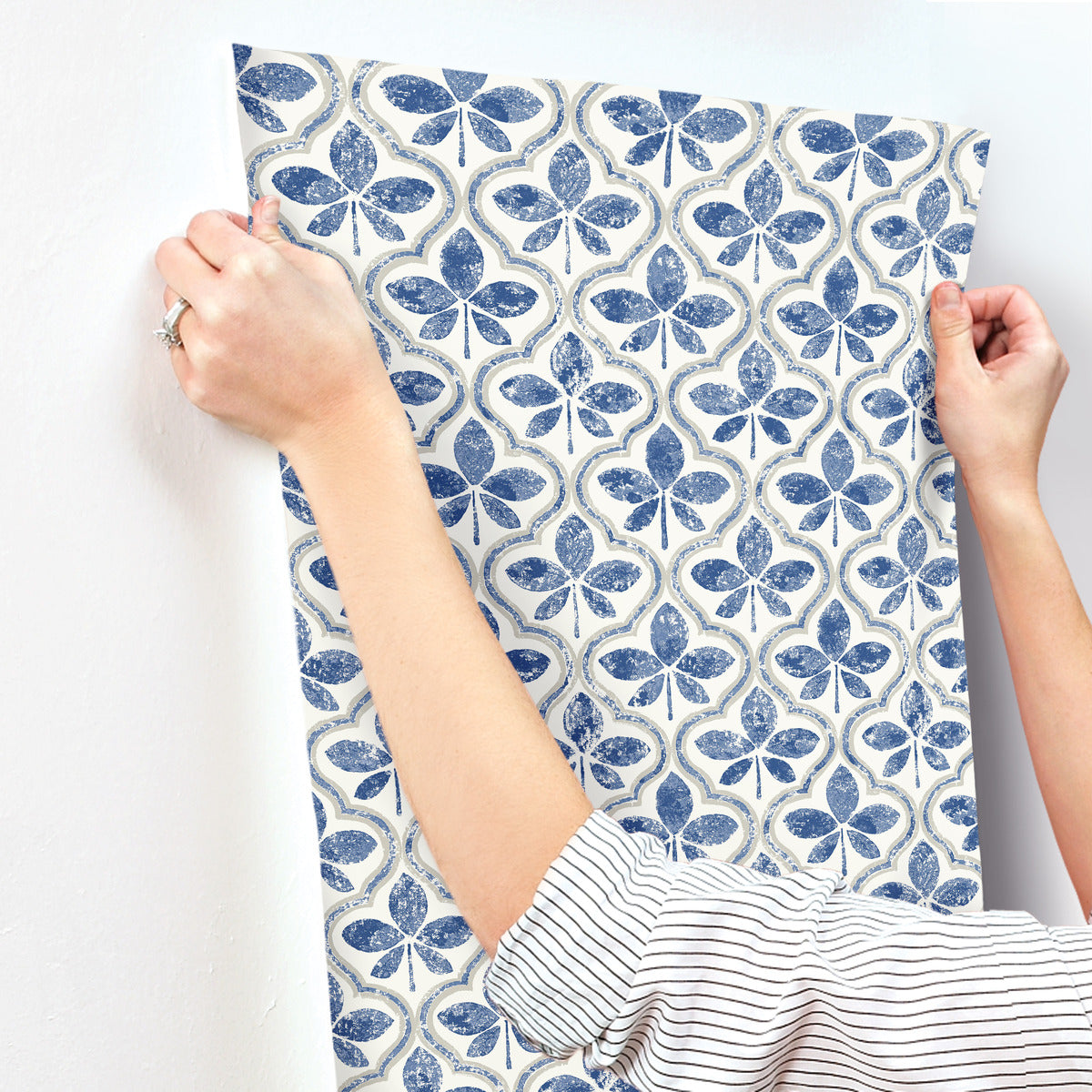 A person with rolled-up sleeves carefully applies York Wallcoverings Sevilla Clover Wallpaper Green (60 Sq.Ft.) with a geometric elegance to a white wall. The blue and white patterned wallpaper features repetitive clover-like shapes. Holding the top corners, their hands smooth it onto the surface using SureStripª technology.