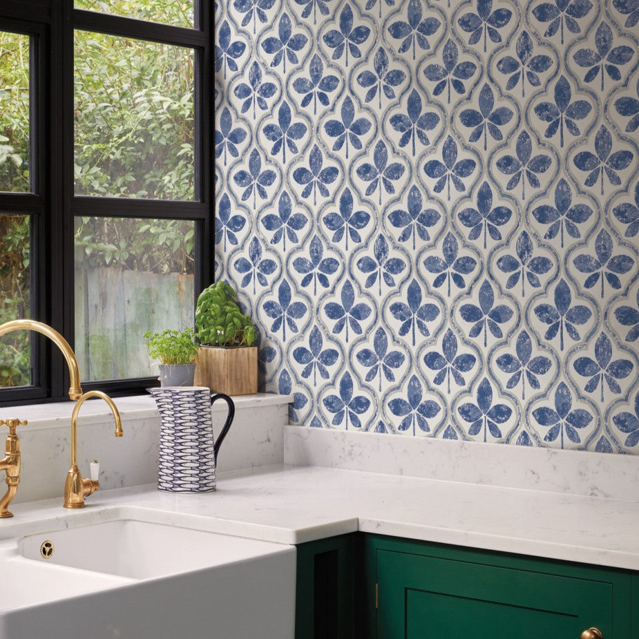 A kitchen with a white and blue patterned York Wallcoverings Sevilla Clover Wallpaper Green (60 Sq.Ft.) exudes geometric elegance. The counter is white marble, featuring a white farmhouse sink with brass fixtures. Near the window, a potted herb plant and striped pitcher add charm, while the lower cabinets are painted green.