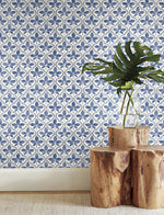 A clear vase with a large green monstera leaf is placed on a stack of three rustic wooden stools of different heights. The background showcases a white wall adorned with York Wallcoverings' Sevilla Onyx Wallpaper Black, Brown (60 Sq.Ft.) featuring an intricate blue floral pattern. The floor is carpeted in a light beige color.