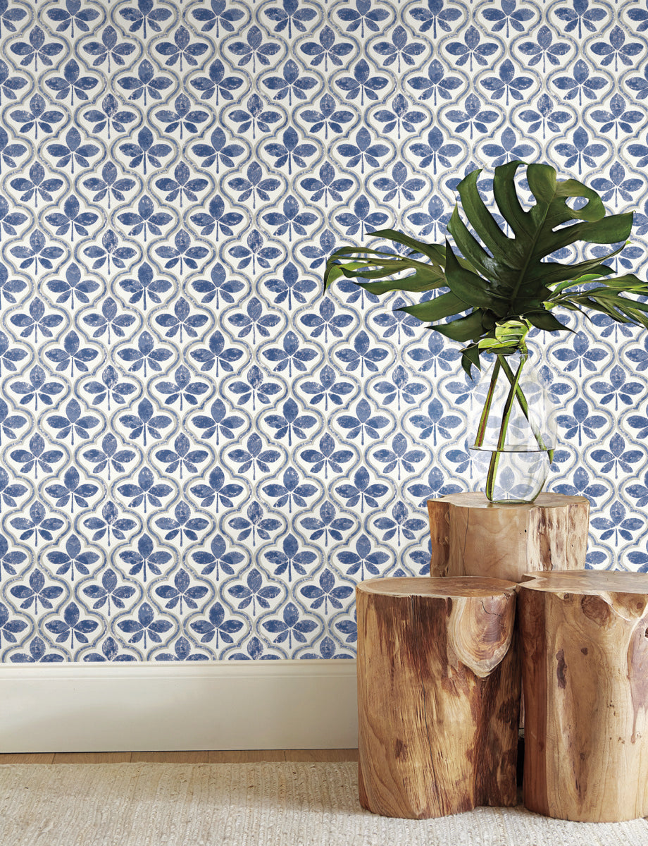 A decorative wall with intricate blue and white tile patterns evokes geometric elegance. In front, a clear glass vase holding a green monstera plant rests on a trio of rustic wooden stump tables. The setting, featuring York Wallcoverings Sevilla Clover Wallpaper Green (60 Sq.Ft.), is completed with a beige carpet and white baseboard.