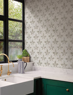 A kitchen corner featuring a white marble countertop with green cabinets. The backsplash showcases the geometric elegance of York Wallcoverings Sevilla Clover Wallpaper Green (60 Sq.Ft.). A white farmhouse sink with a gold faucet is installed beside a window with black framing. A plant and jug are on the countertop.